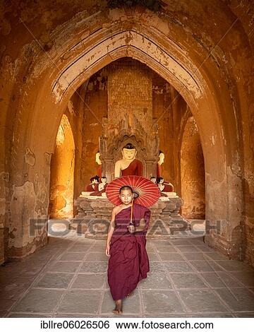 Stock Photograph - Buddhist young monk in red robe with red umbrella walks in a temple, Bagan, Myanmar, Asia. Fotosearch