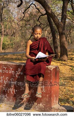 Stock Photograph - Child monk with red robe reading on a red wall, Shwe Hin Thar Monastery, Sagaing, Myanmar, Asia. Fotosearch