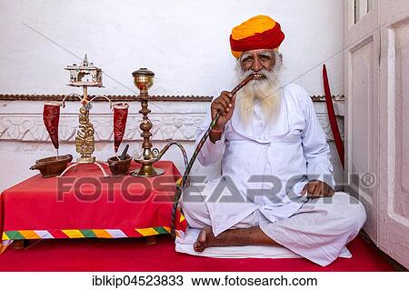 Stock Image - Elderly Indian man with turban smoke hookah, Mehrangarh Fort, Jodhpur, Rajasthan, India, Asia. Fotosearch