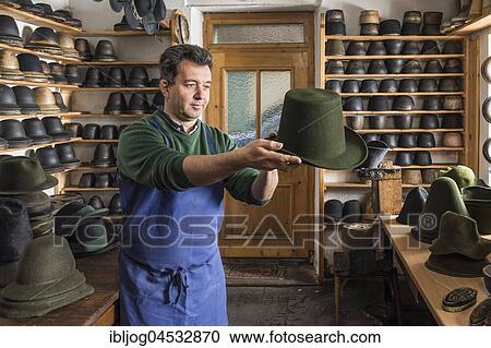 Stock Image - Hatter examining finished wool felt hat, hat molds on wooden shelves and finished hats behind, hatmaker workshop, Bad Aussee, Styria, Austria, Europe. Fotosearch