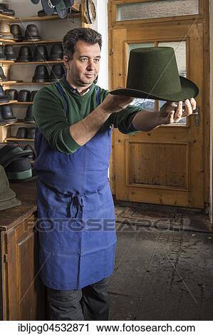 Stock Image - Hatter examining finished wool felt hat, hat molds on wooden shelves and finished hats behind, hatmaker workshop, Bad Aussee, Styria, Austria, Europe. Fotosearch