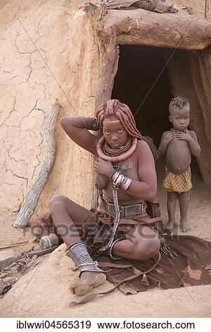 Stock Photo - Himba woman with child in front of the mud hut, rubs itself with Okra, butterfat with ochre paint, Kaokoveld, Namibia, Africa. Fotosearch