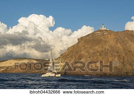Stock Photograph - Sailboat passing lighthouse, Akrotiri, Santorini, Cyclades, Greece, Europe. Fotosearch