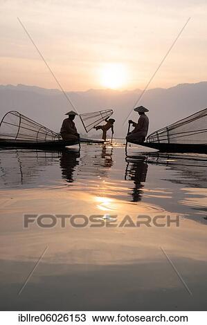 Stock Image - Three traditional fishermen pose standing and sitting on their small boats in front of the sun, Lake Inle, Myanmar, Asia. Fotosearch