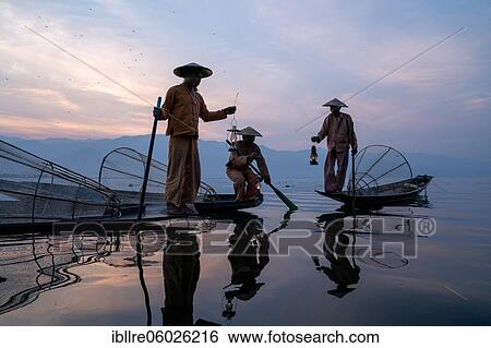 Stock Photograph - Three traditional fishermen sit and stand with lamps on their small boats, at sunrise, Lake Inle, Myanmar, Asia. Fotosearch