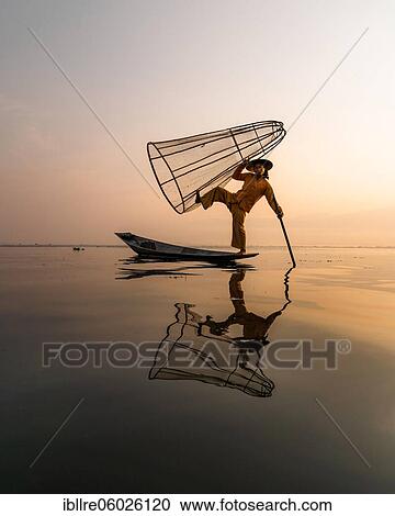 Stock Image - Traditional fisherman posing standing on his small boat with reflection in front of sunrise, Lake Inle, Myanmar, Asia. Fotosearch