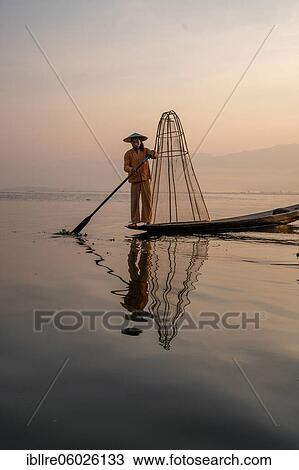 Stock Image - Traditional fisherman posing standing on his small boat with reflection in front of sunrise, Lake Inle, Myanmar, Asia. Fotosearch