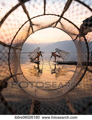 Stock Image - Two traditional fishermen pose on their small boats, photographically framed by a fishing net at sunrise, Lake Inle, Myanmar, Asia. Fotosearch