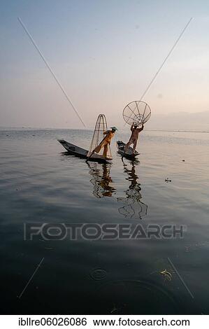 Stock Photograph - Two traditional fishermen pose standing on their small boats, at sunrise, Lake Inle, Myanmar, Asia. Fotosearch