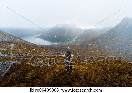Stock Photo - Woman looking at the village Fjordbotn and a prominent and pointed mountain, snowfall in autumnal fjord landscape, Fjordgard, Senja, Norway, Europe. Fotosearch