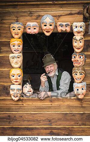 Stock Image - Wooden mask carver in a window, framed by painted wooden masks, wooden mask carving workshop, Bad Aussee, Styria, Austria, Europe. Fotosearch