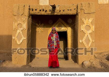 Stock Image - Young girl in traditional clothing in front of a mud house, Agadez, Niger, Africa. Fotosearch