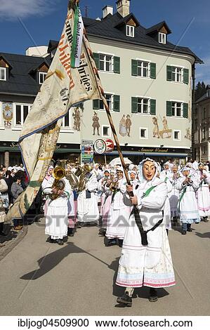 Stock Image - Aussee Carnival, man with mask swinging flag, at back men with masks and instruments, white robes, Trommelweiber, Bad Aussee, Styria, Austria, Europe. Fotosearch