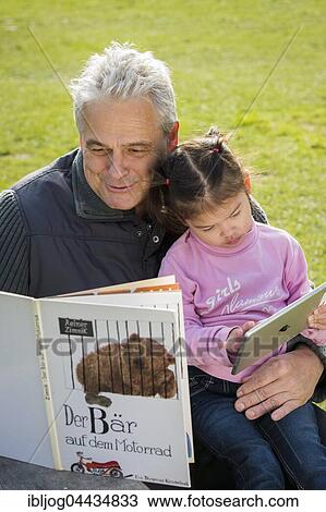 Stock Image - Father reading book to daughter, sitting on meadow, girl playing with tablet, Poing, Bavaria, Germany, Europe. Fotosearch