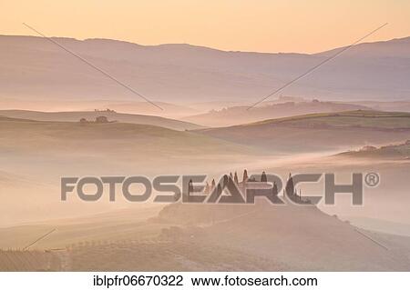Stock Image - Hilly landscape with cornfields and cypresses, manor house, group of cypresses, traditional Tuscany stone house, fog, morning atmosphere, in spring, Tuscany, Italy, Europe. Fotosearch
