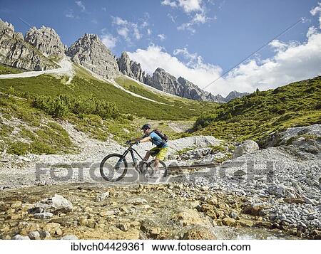 Stock Image - Mountainbiker rides with his mountain bike through a stream, in the background mountain chain Kalkkögel, Stubai Alps, Tyrol, Austria, Europe. Fotosearch
