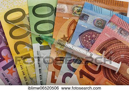 Stock Photograph - Symbolic picture Costs of mass vaccination against Covid-19, syringe lies on EURO banknotes, corona crisis, Baden-Württemberg, Germany, Europe. Fotosearch
