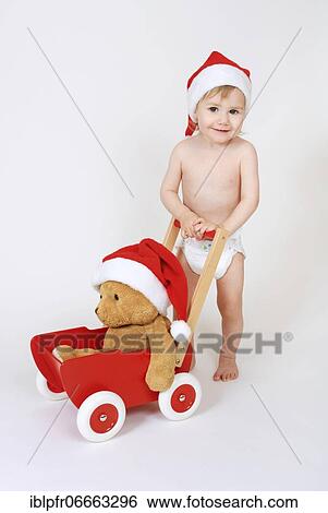 Stock Photograph - Toddler standing, playing with teddy bear and wearing christmas cap. Fotosearch