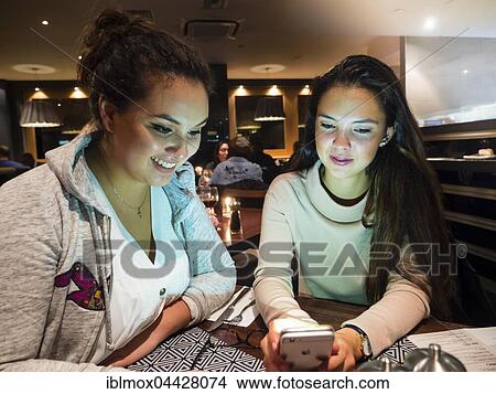 Picture - Two young women with mobile phone in a restaurant, London, England, United Kingdom, Europe. Fotosearch