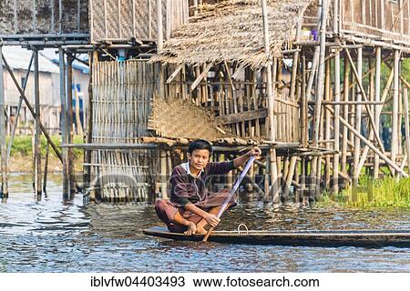 Stock Image - Boy, young man rowing, traditional stilt houses behind, Inle Lake, Nampan, Shan State, Myanmar, Asia. Fotosearch