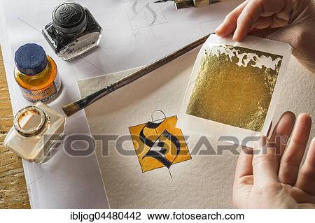 Stock Image - Calligraphy studio, hands holding sheet with transfer gold, letters S on torchon paper at back, inkwells, paintbrush, Seebruck, Upper Bavaria, Germany, Europe. Fotosearch