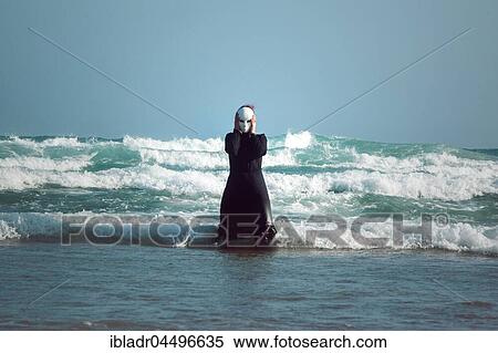Stock Photography - Woman in black dress with mask, standing in the surf in the sea. Fotosearch
