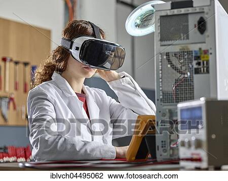 Stock Image - Woman with VR goggles and lab coat sitting in front of PC open in an electronics laboratory, Austria, Europe. Fotosearch