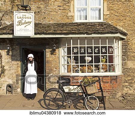 Stock Photo - Young shopkeeper woman in old fashioned traditional clothing outside the village bakery shop, Lacock, Wiltshire, England, UK. Fotosearch