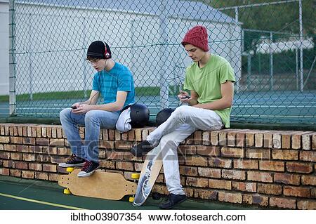 Picture - Two teenage boys sitting on a brick wall, with skateboards, listening to music and using a cell phone. Fotosearch