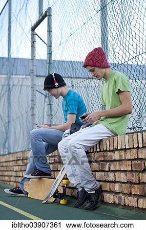 Two teenage boys sitting on a brick wall, with skateboards, listening to music and using a cell phone View Large Photo Image Stock Image - Two teenage boys sitting on a brick wall, with skateboards, listening to music and using a cell phone. Fotosearch