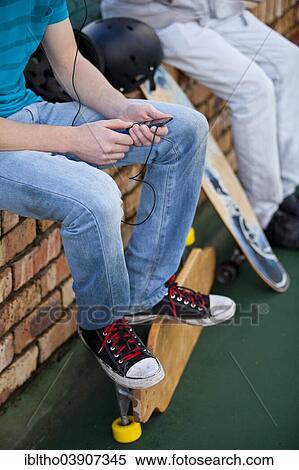 Stock Photography - Two teenage boys sitting on a brick wall, with skateboards, listening to music. Fotosearch
