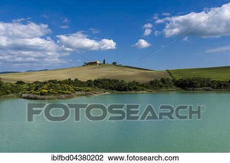 Stock Image - Typical green Tuscan landscape in Val d’Orcia, farm on hill, lake and blue cloudy sky, La Foce, Tuscany, Italy, Europe. Fotosearch