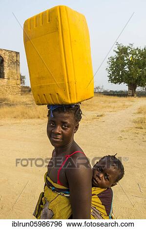 Stock Photograph - Woman with her baby on the back carrying a water canister on her head, Massangano, Cuanza Norte, Angola, Africa. Fotosearch