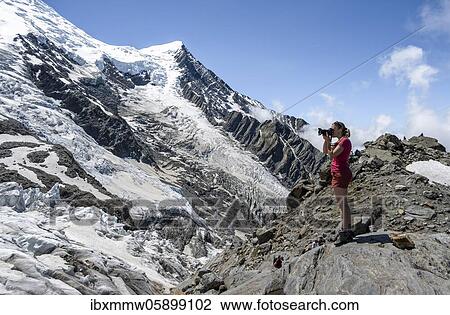 Stock Image - La Jonction, hiker taking photos of glacier tongue, Glacier des Bossons meets Glacier de Taconnaz, left summit Mont Maudit, right Mont Blanc, Chamonix, Haute-Savoie, France, Europe. Fotosearch