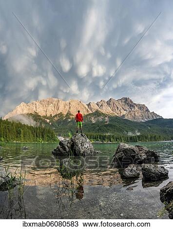 Stock Image - Young man standing on a rock on the shore, Eibsee lake in front of Zugspitze massif with Zugspitze, sunset, dramatic Mammaten clouds, Wetterstein range, near Grainau, Upper Bavaria, Bavaria, Germany, Europe. Fotosearch