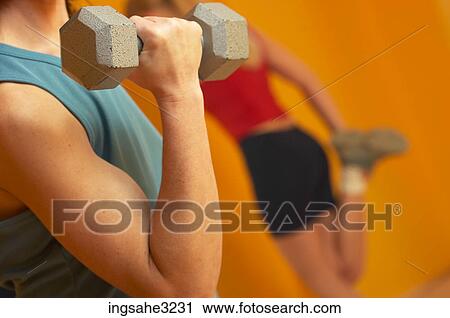 Stock Image - Mature adult woman lifting weights with woman stretching in background . Fotosearch