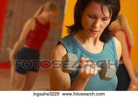 Stock Photo - Mature adult woman lifting weights with woman stretching in background . Fotosearch