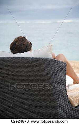Stock Photograph - Back view of a woman sitting on a chaise chair on beach in Bali. Fotosearch