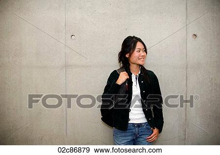 Stock Photo - Smiling Asian student leaning on school cement wall.. Fotosearch