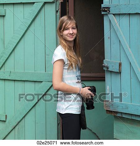 Stock Image - Teenage girl standing between wooden doors holding a camera. . Fotosearch