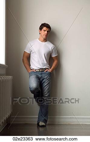 Stock Image - Young adult man leaning against white wall, wearing a white t-shirt.. Fotosearch