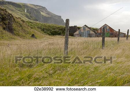 Stock Image - Barbwire fence and fenceposts in a pasture with barns and mountains. Fotosearch