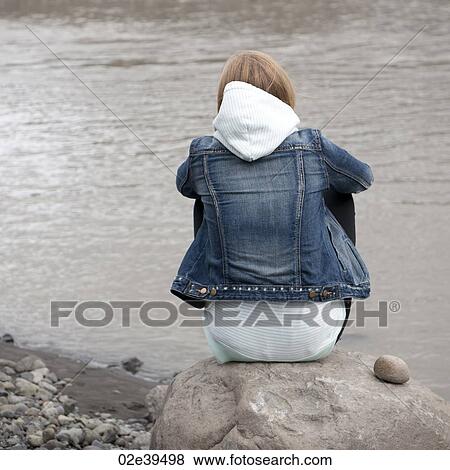 Girl sitting on rock at lakeshore in denim jacket and hooded pullover View Large Photo Image Stock Photo - Girl sitting on rock at lakeshore in denim jacket and hooded pullover. Fotosearch