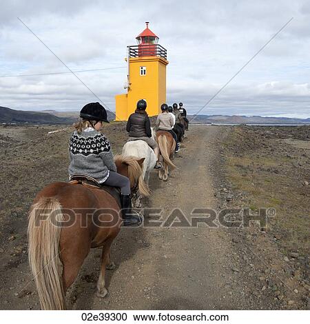 Stock Image - Group of people riding Icelandic horses along a path to lighthouse. Fotosearch