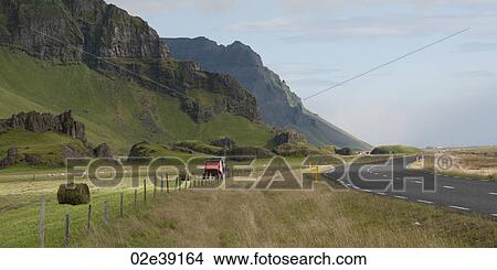 Picture - Hay baler making bales on farmland between mountainside and highway. Fotosearch