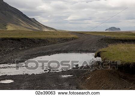 Stock Image - Landscape of mountain and pasture with rural dirt road and puddles. Fotosearch