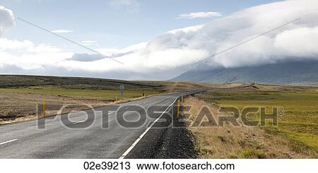 Stock Image - Paved highway disappearing into horizon towards clod covered mountains. Fotosearch