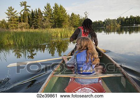 Stock Image - Rear view of blond girl and Africian American man rowing in a canoe at Lake of the Woods, Ontario. Fotosearch
