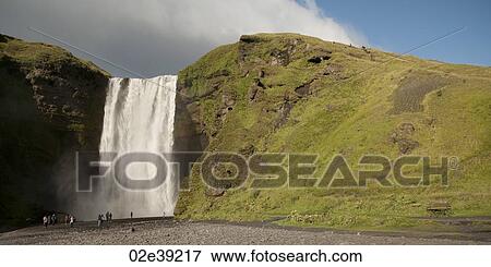 Stock Photo - Skagafoss waterfall over grassy cliffside with sightseers. Fotosearch