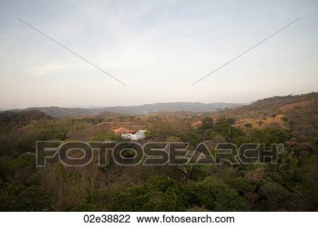 Stock Image - Sun fading over a white house with an orange roof in the rolling hills above the treetops of Costa Rica. Fotosearch
